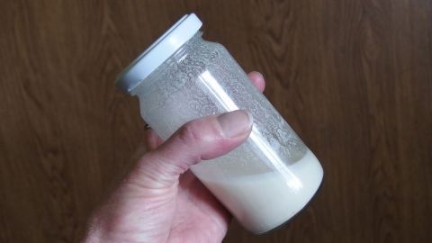 Ein Glas mit einer hellen, cremigen Flüssigkeit wird von einer Hand gehalten, um eine Klümpchen-freie Mehlschwitze zuzubereiten.