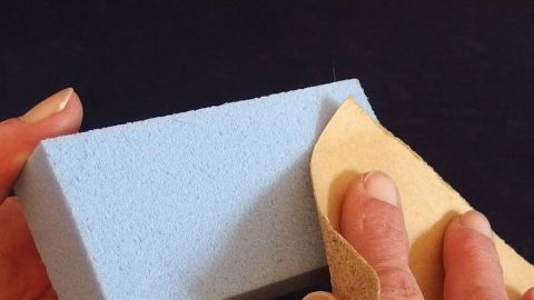 Ein blauer Bimsstein wird mit einem hellen Schwamm gereinigt, während die Hände ihn in einem dunklen Umfeld halten.