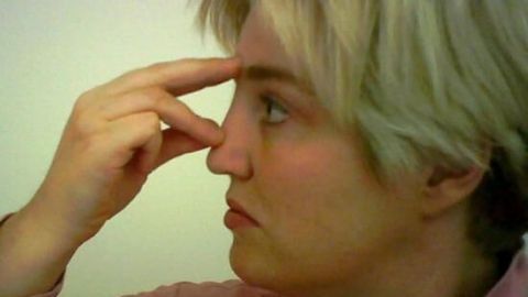 Eine Person drückt mit einem Finger auf die Nase, um die Atmung zu erleichtern. Der Hintergrund ist neutral.