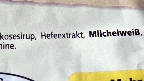 Die Verpackung zeigt Inhaltsstoffe, darunter "Milcheiweiß", während Verbraucher über Geschmacksverstärker informiert werden.