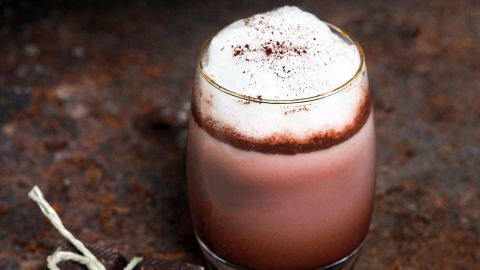 Ein schaumiger Cappuccino in einem transparenten Glas steht neben einem Stück Schokolade auf einer rustikalen Oberfläche.