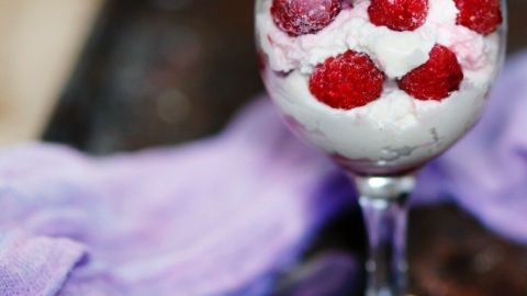 Ein Dessertglas gefüllt mit Sahne, Himbeeren und rosa Baisers steht auf einem rustikalen Tisch, umgeben von lila Stoff.
