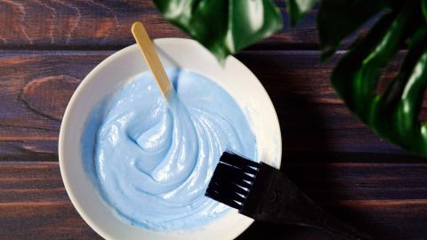 Eine Schüssel mit blauer Pflanzenfarbe steht auf einem Holztisch, umgeben von grünen Blättern. Ein Pinsel liegt bereit.