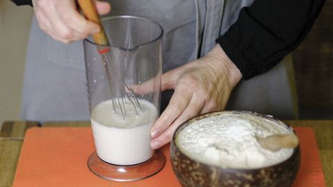 Eine Hand schlagt Sahne in einem Becher mit einem Schneebesen, während eine Schüssel mit Mehl auf der Arbeitsfläche steht.