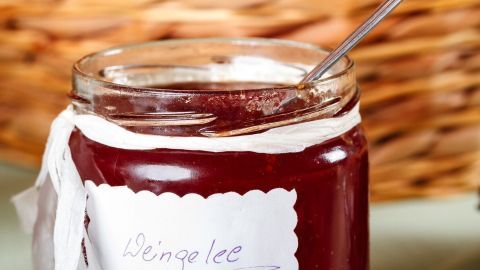 Ein Glas Weingelee steht auf dem Tisch, ein Löffel steckt darin, umgeben von einem geflochtenen Körbchen im Hintergrund.