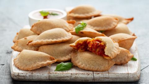 Kärntner Nudeln liegen auf einem Holzbrett, einige sind aufgebrochen, und zeigen eine Füllung mit Tomate-Mozzarella.