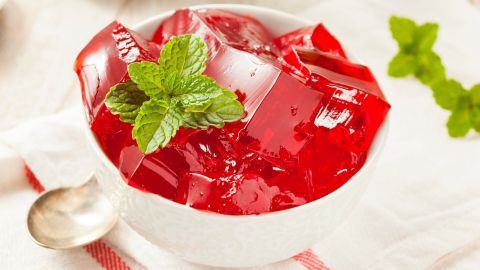 Ein Glas Götterspeise mit roten Wackelstücken und frischen Minzblättern steht in einer weißen Schüssel auf einem Tuch.