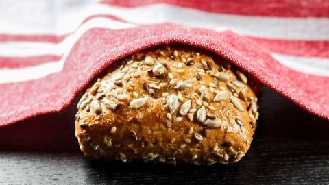 Ein goldbraunes Brötchen mit Saaten liegt unter einem rot-weißen Geschirrtuch auf einem dunklen Tisch.