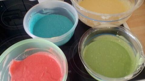 Bunte Kuchenteige in Gelb, Blau, Rot und Grün stehen in Schüsseln auf einem Herd, bereit für einen Papageienkuchen.