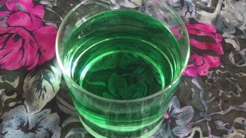 Ein Glas mit grüner Flüssigkeit steht auf einem blühenden, bunten Tischmuster und wirkt einladend und verspielt.