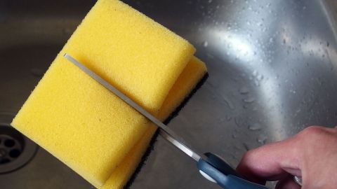 In der Mitte halbierte Topfschwämme sind handlicher und außerdem hat man doppelt so viel Schwämme für gleiches Geld. Ein gelber Topfschwamm wird in einer Spüle mit einer Schere halbiert, um ihn handlicher zu machen.
