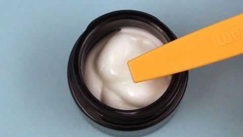Eine schwarze Creme-Packung zeigt eine weiße, cremige Textur, während ein gelber Spatel die Creme entnimmt, um weniger Pickel zu fördern.