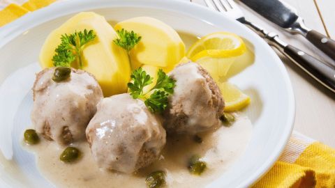 Königsberger Klopse liegen in einer sahnigen Soße, ergänzt durch Kartoffeln und Zitronenscheiben auf einem weißen Teller.