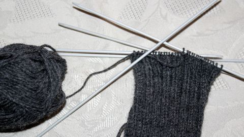 Dunkles Garn und Stricknadeln liegen auf einem Tisch, während ein Ärmel in Ribbelmuster langsam entsteht.