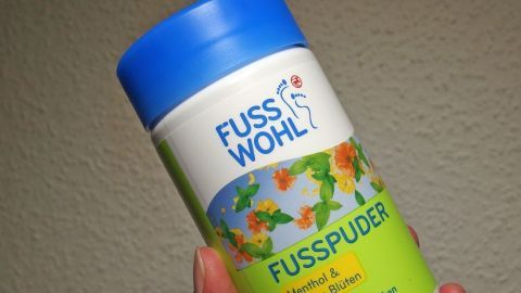 Ein Hand hält ein farbenfrohes Verpackung von Fußpuder, das Insekten vertreibt, vor einer neutralen Wand.
