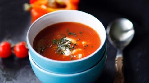 Eine tiefblaue Schüssel mit Tomaten-Paprikasuppe steht auf einem dunklen Hintergrund, umgeben von roten Paprikastücken und Kirschtomaten.