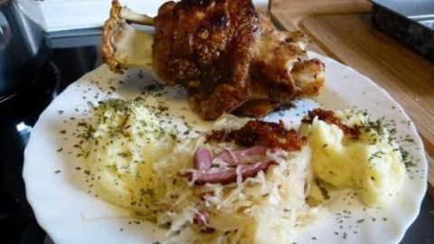 Eine knusprige Schweinshaxe liegt auf einem Teller, umgeben von Kartoffeln und Sauerkraut, appetitlich angerichtet.