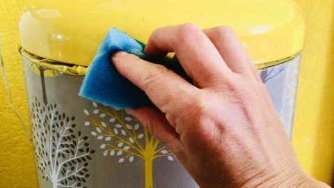 Ein Küchenmülleimer wird mit einem blauen Schwamm gereinigt, vor einer gelben Wand im Hintergrund.