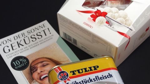 Ein buntes Sortiment liegt auf einem Tisch: eine Werbebroschüre mit „10% Rabatt“, ein festlich verpackter Kuchen und eine Dose Frühstücksfleisch.