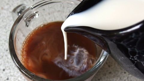 Eine Kanne gießt Milch in ein Glas mit starkem, kalten Eiskaffee auf einer hellen Oberfläche.