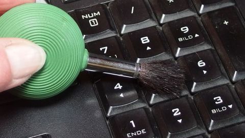 Ein kleiner, grüner Blasebalg mit Pinsel reinigt ein Keyboard, während eine Hand das Gerät führt.