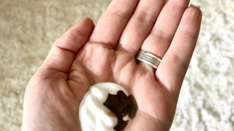 Eine Hand hält eine Mischung aus Pflegecreme und Selbstbräunungscreme, umgeben von einem hellen, flauschigen Hintergrund.