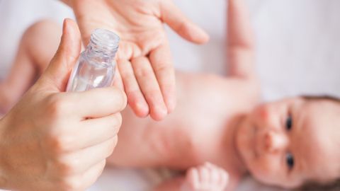 Eine Hand hält eine Flasche, während eine weitere Hand Babyöl auf die andere Hand aufträgt, um das Baby zu massieren.