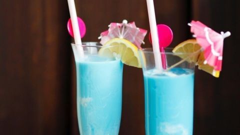 Zwei glitzernde Cocktails in Flöten mit blauer Farbe, Schirmchen und Früchten, stehen auf einem Holztisch vor dunklem Hintergrund.