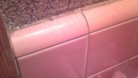 Nagelfeile liegt auf pinken Fliesen in einem Badezimmer neben einer kleinen Parfümflasche; rustikale Wand als Hintergrund.