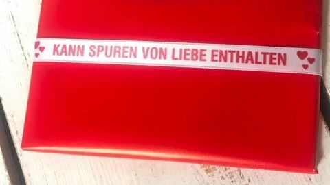 Ein rotes Geschenk ist liebevoll verpackt und liegt auf einem weißen Holztisch. Die Schleife trägt den Text: "KANN SPUREN VON LIEBE ENTHALTEN".