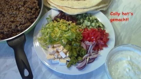 Eine Pfanne mit Hackfleisch liegt neben einer Platte mit frischen Zutaten für Pseudo-Döner und Fladenbrot auf einem Tisch.