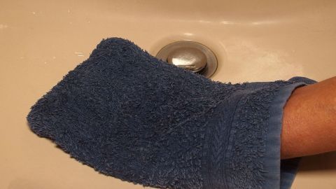 Ein blauer Waschlappen wird verwendet, um einen Waschbeckenabfluss zu reinigen, während eine Hand damit arbeitet.