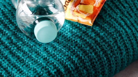 Ein grünes Wollkleidungsstück liegt gestapelt mit einer Flasche Mineralwasser und einem Päckchen Backpulver daneben.