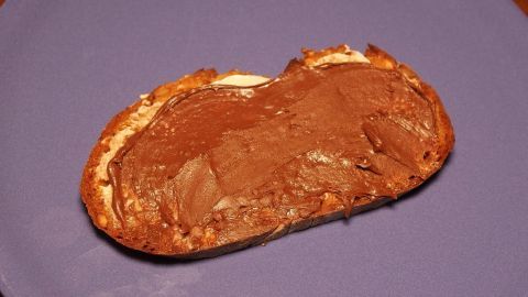 Aus Margarine, Kakaopulver und gemahlenen Haselnüssen kann man einen nutellaähnlichen Brotaufstrich selber machen. Ein Stück Brot belegt mit hausgemachter Nutella liegt auf einem blauen Teller. Der Schokoladenaufstrich glänzt appetitlich.