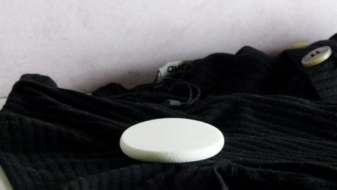 Ein runder, weißer Fleckenentferner liegt auf einem schwarzen Kleidungsstück auf einem Tisch vor einer hellen Wand.