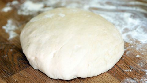 Ein runder Hefeteig liegt auf einer bemehlten Holzfläche und wartet darauf, weiterverarbeitet zu werden.