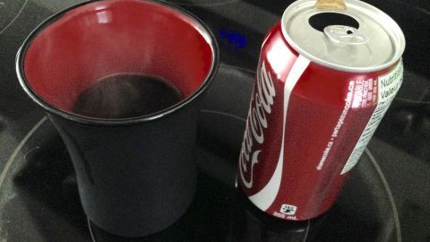 Ein schwarzer Becher mit warmer Cola steht neben einer geöffneten Cola-Dose auf einer Kochplatte.