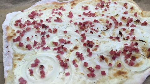 Ein frisch gebackener Elsässer Flammkuchen mit Hefeteig, belegt mit Quark und Speck, liegt auf einem Backblech.