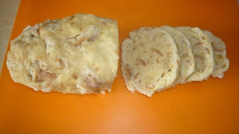 Ein geformtes Stück Böhmischer Knödel liegt auf einem orangefarbenen Schneidebrett, daneben befinden sich gleichmäßige Scheiben.