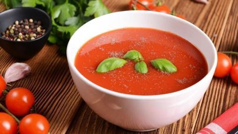 Tomatensuppe in weißer Schale, garniert mit Basilikum, umgeben von Tomaten, Pfeffer und Kräutern auf Holztisch.