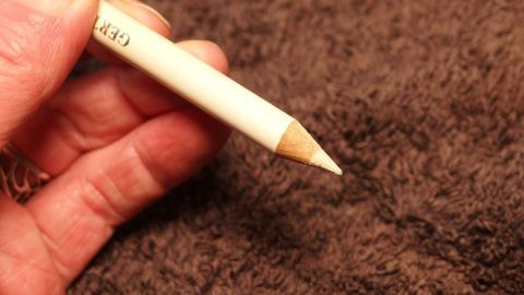 Ein weißer Kajalstift wird von einer Hand gehalten, umgeben von einem braunen Hintergrund. Stift beschriftet mit "GERX".