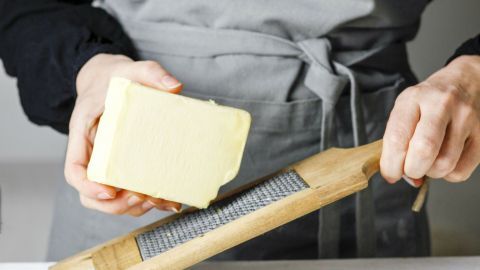 Ein Stück Butter wird auf einer Reibe gehalten und darüber gerieben, um sie weicher zu machen; der Hintergrund ist hell und neutral.