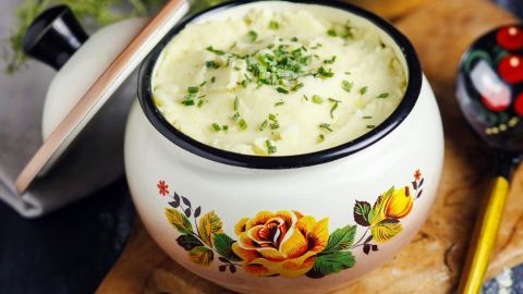 Ein kunstvoll verziertes Schüsselchen mit cremigem Kartoffelbrei, garniert mit frischen Kräutern, auf einem Holzbrett.