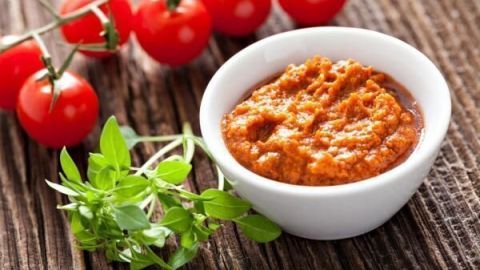 Tomatenpesto in weißer Schale, dekoriert mit frischen Kräutern, daneben liegen frische Kirschtomaten auf rustikalem Tisch.