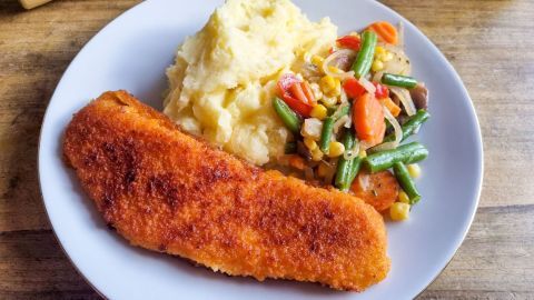 Fettarmes, hauchdünnes paniertes Filet liegt neben cremigen Kartoffelpüree und buntem Gemüse auf einem weißen Teller.