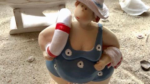 Eine Figur einer runden Frau trägt einen blauen Badeanzug und hält einen Rettungsring, umgeben von Muscheln.