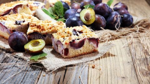 Ein leckerer Pflaumen-Streuselkuchen liegt auf einem rustikalen Holztablett, umgeben von frischen Pflaumen und grünem Blattwerk.
