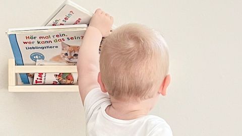 Ein kleiner Junge in einem weißen T-Shirt greift nach einem Buch in einem Bücherregal an einer Wand.