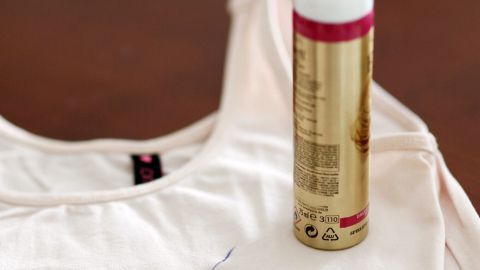 Ein Haarspray steht senkrecht auf einem hellen T-Shirt, daneben liegt ein blauer Stift. Es wird gezeigt, wie man Haarspray gegen abfärbende Schuhe nutzen kann.