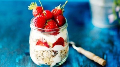 Ein Schraubglas enthält ein Müsligemisch mit Joghurt und frischen Erdbeeren, auf einem leuchtend blauen Tisch, daneben steht eine Blumenvase.